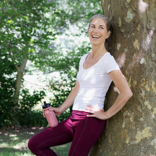 Frau in Sport-Outfit lehnt lächelnd an Baum mit pinker Trinkflasche – EverGlow Shake Wohlfühl-Porträt
