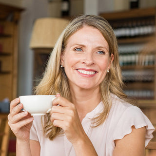 Blonde Frau lächelt und hält eine weisse Teetasse in der Hand vor warmem Hintergrund